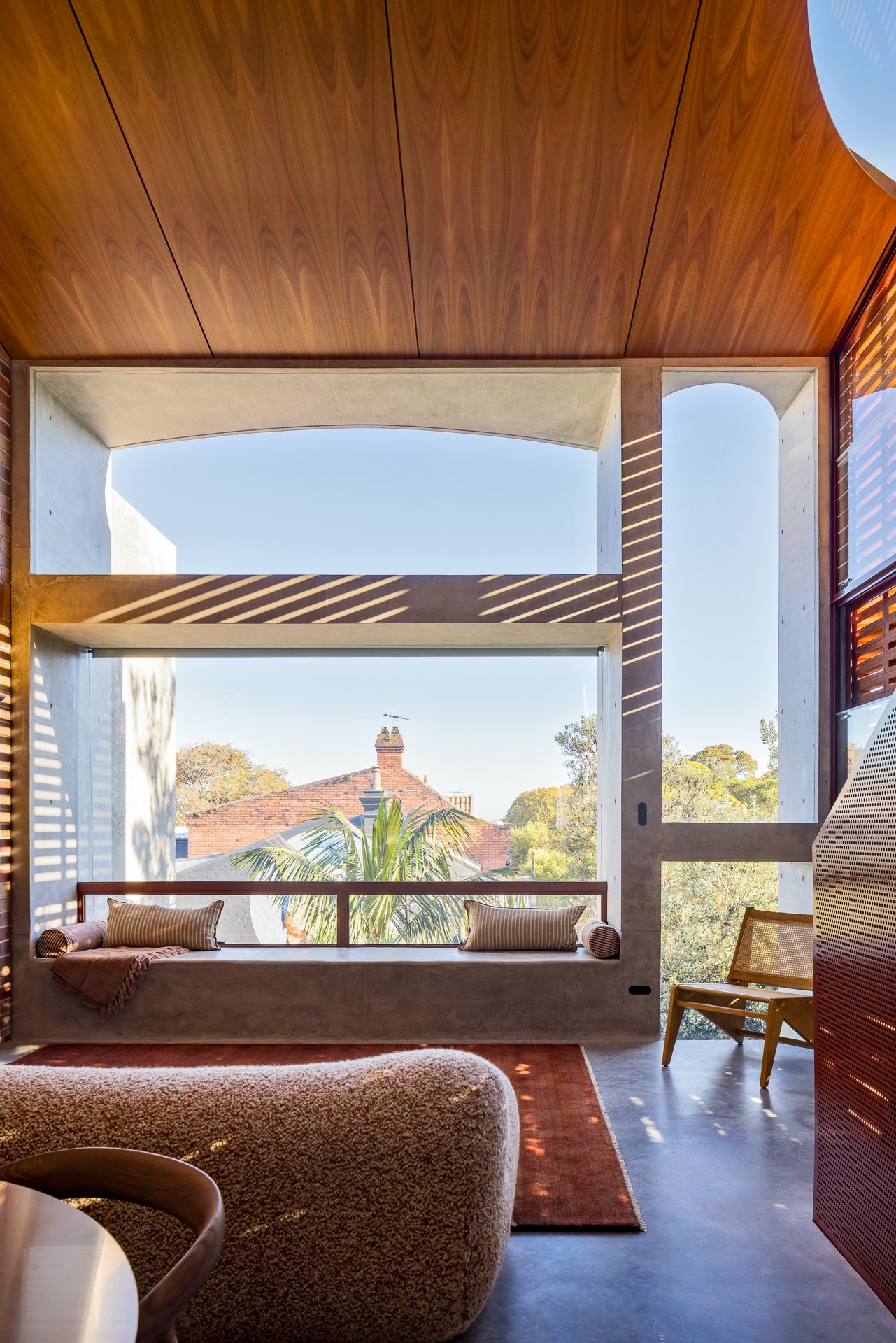 Built-in window seat with cushions beside a large window with timber ceiling, overlooking trees and neighboring rooftops.