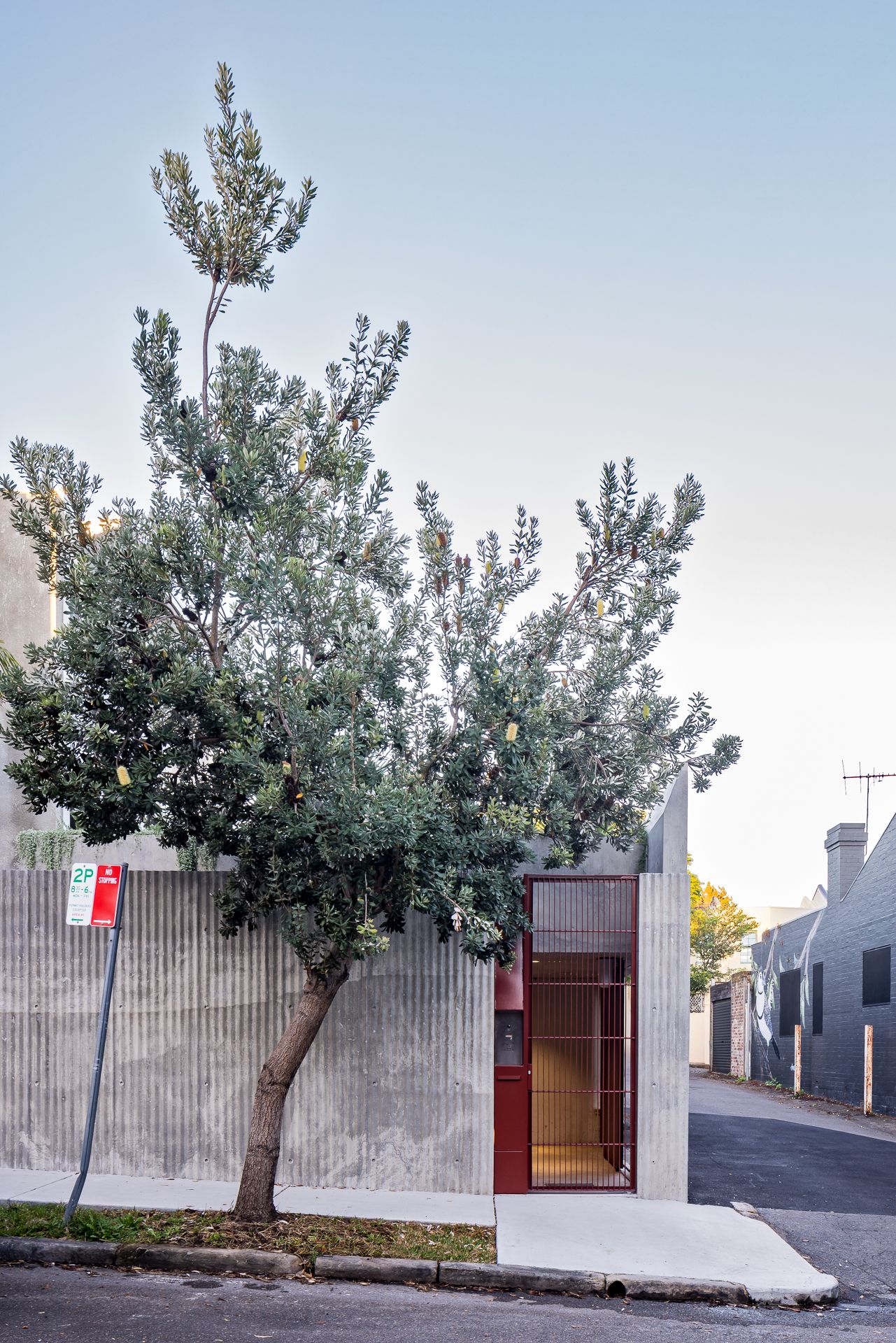 Narrow concrete and brick townhouse facade with a red entry gate and a leafy tree on a quiet street corner at dusk.