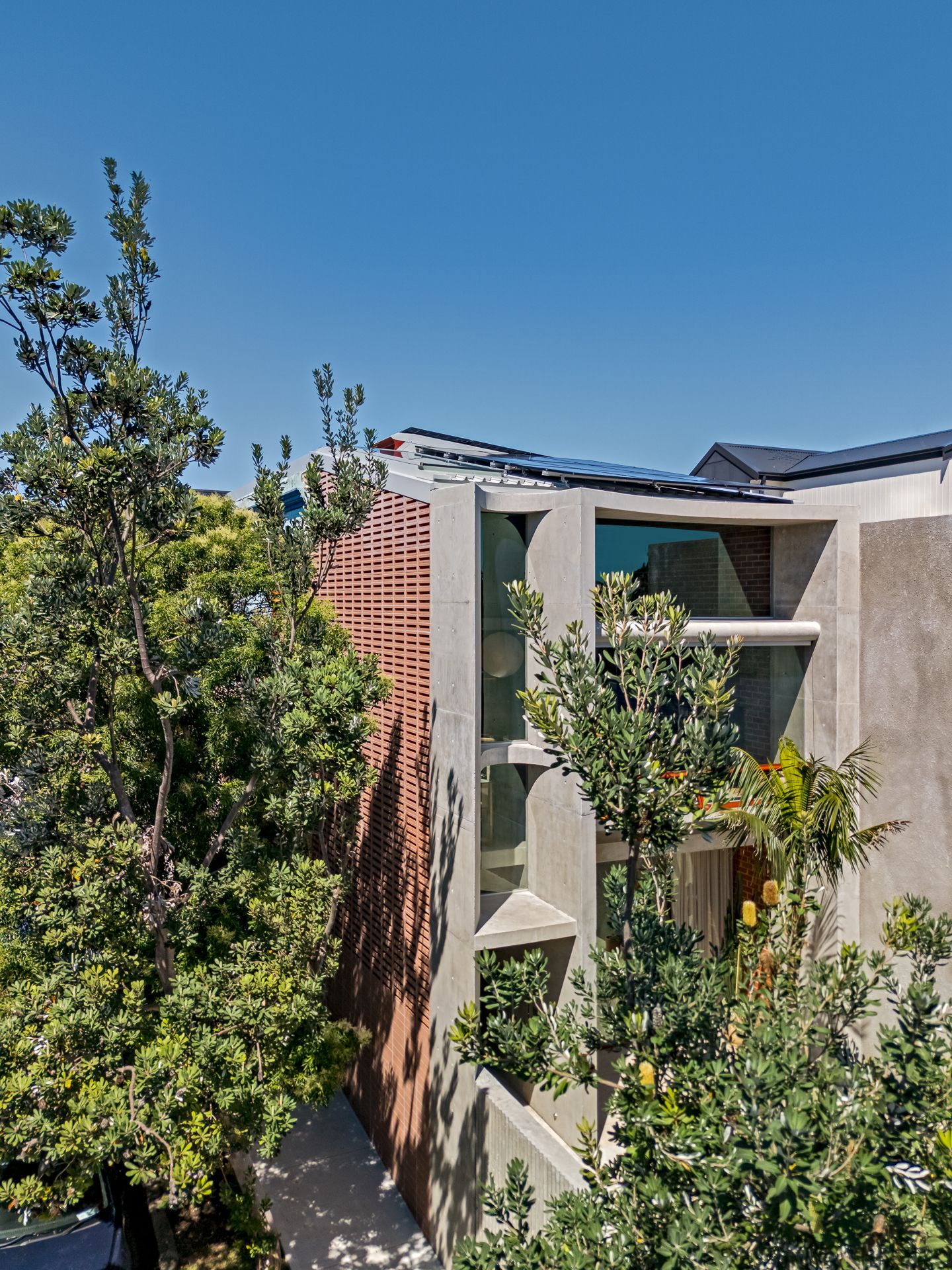 Modern concrete and brick residential building with large windows and rooftop solar panels surrounded by lush green trees.