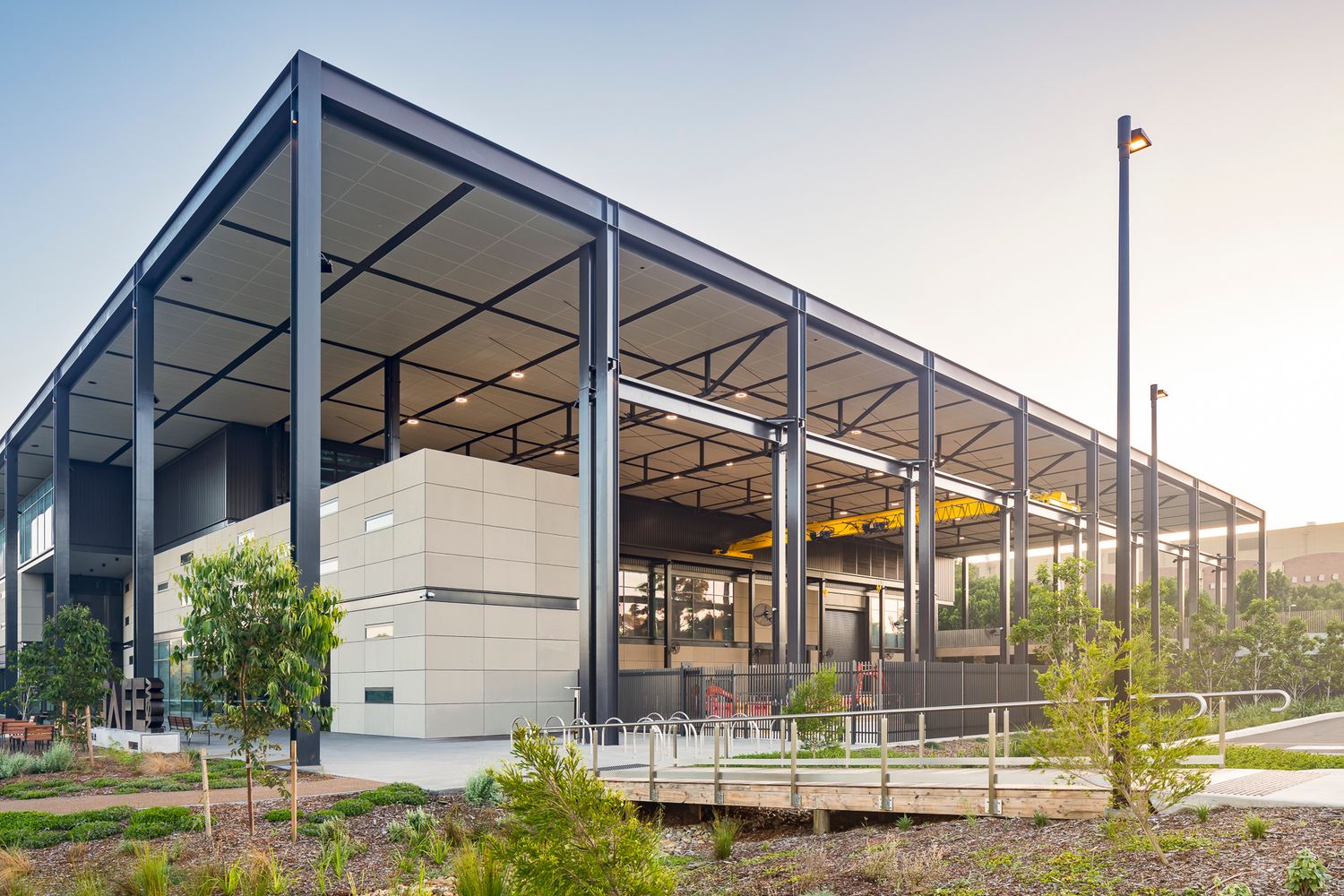 Modern steel-framed building at twilight with a wide roof overhang, warm interior lighting, native gardens and a timber boardwalk.