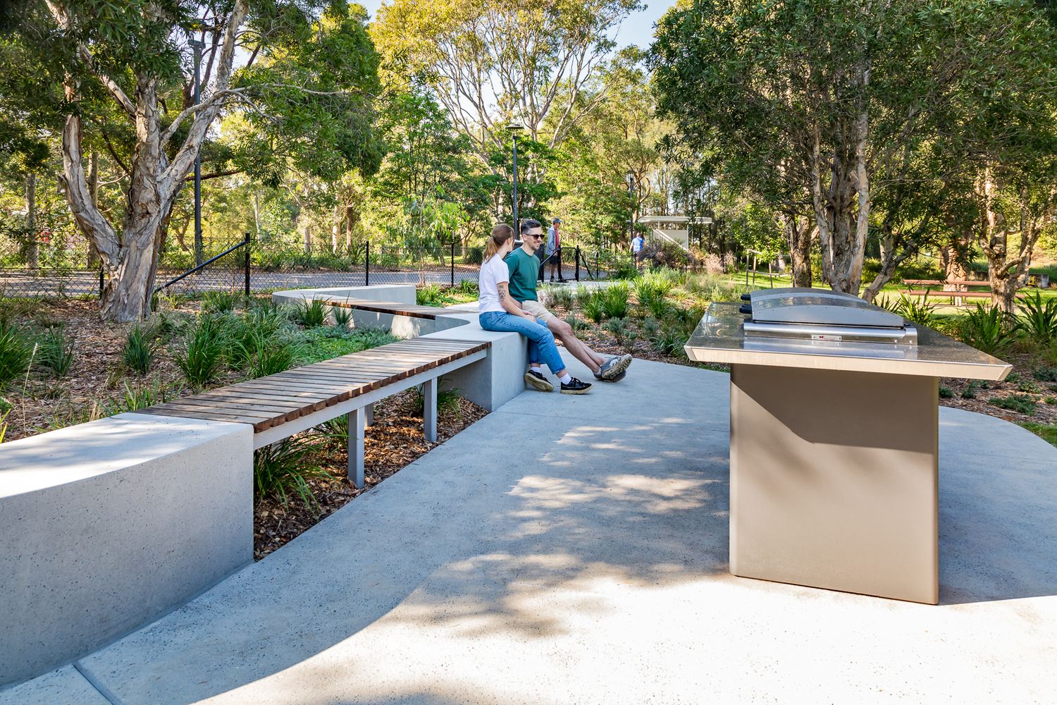 Curved concrete pathway with built-in benches and picnic tables winds through native plantings and gum trees in a shady park.