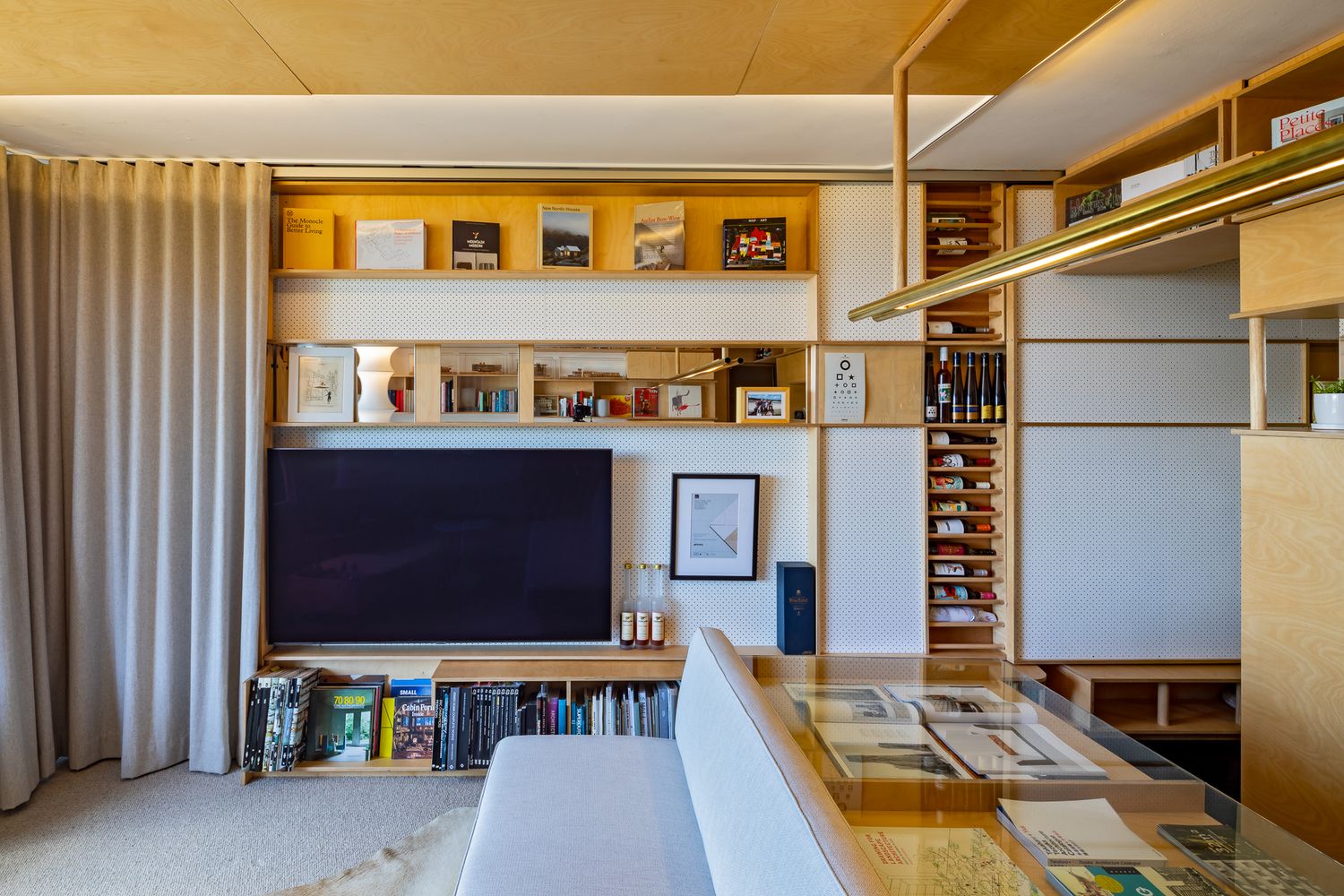 Modern living room with a wall-mounted TV, wooden shelving, books and framed art beside a beige curtain.