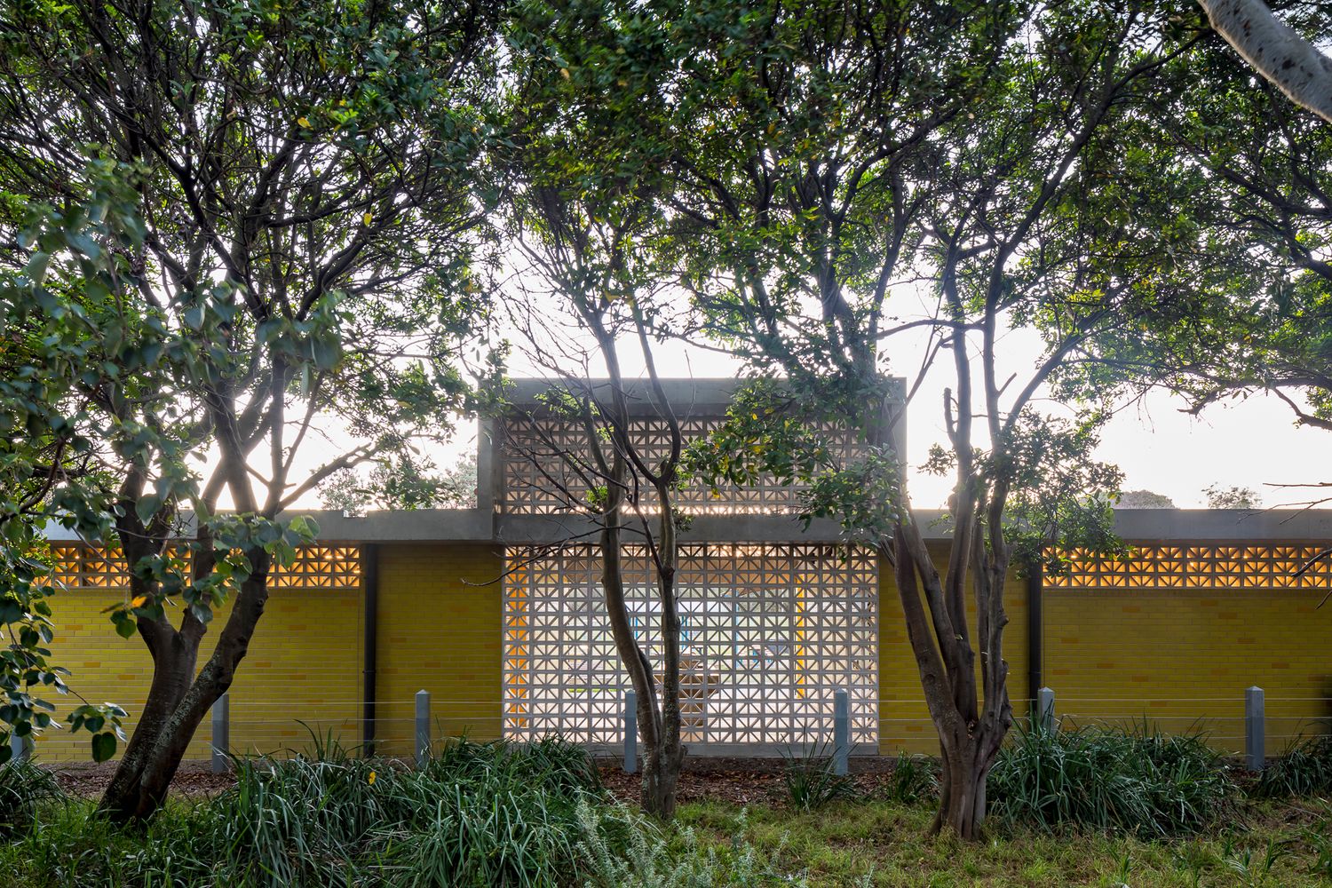 Amenities building with yellow brick walls and white breeze block screens seen through leafy trees and native coastal plants.