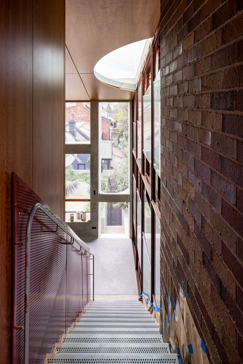 Narrow interior stairwell with warm timber panelling, a textured brick wall, perforated metal balustrade and a curved skylight above.
