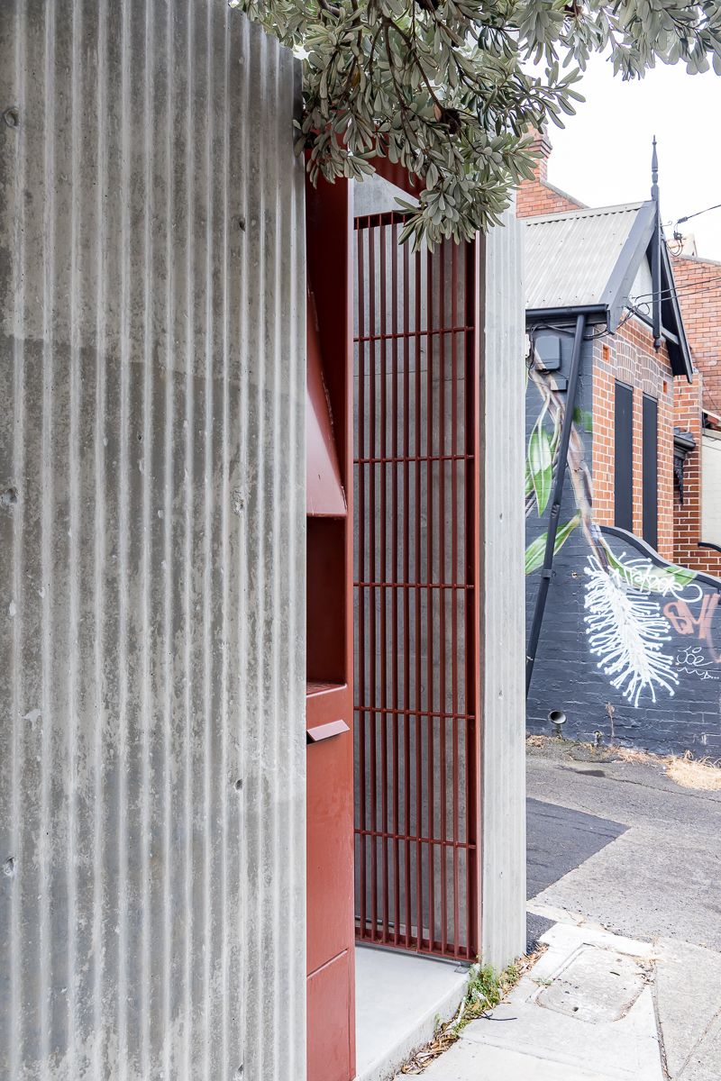 Corrugated concrete wall beside a red metal slatted gate on a laneway, with a street mural and brick buildings in the background.