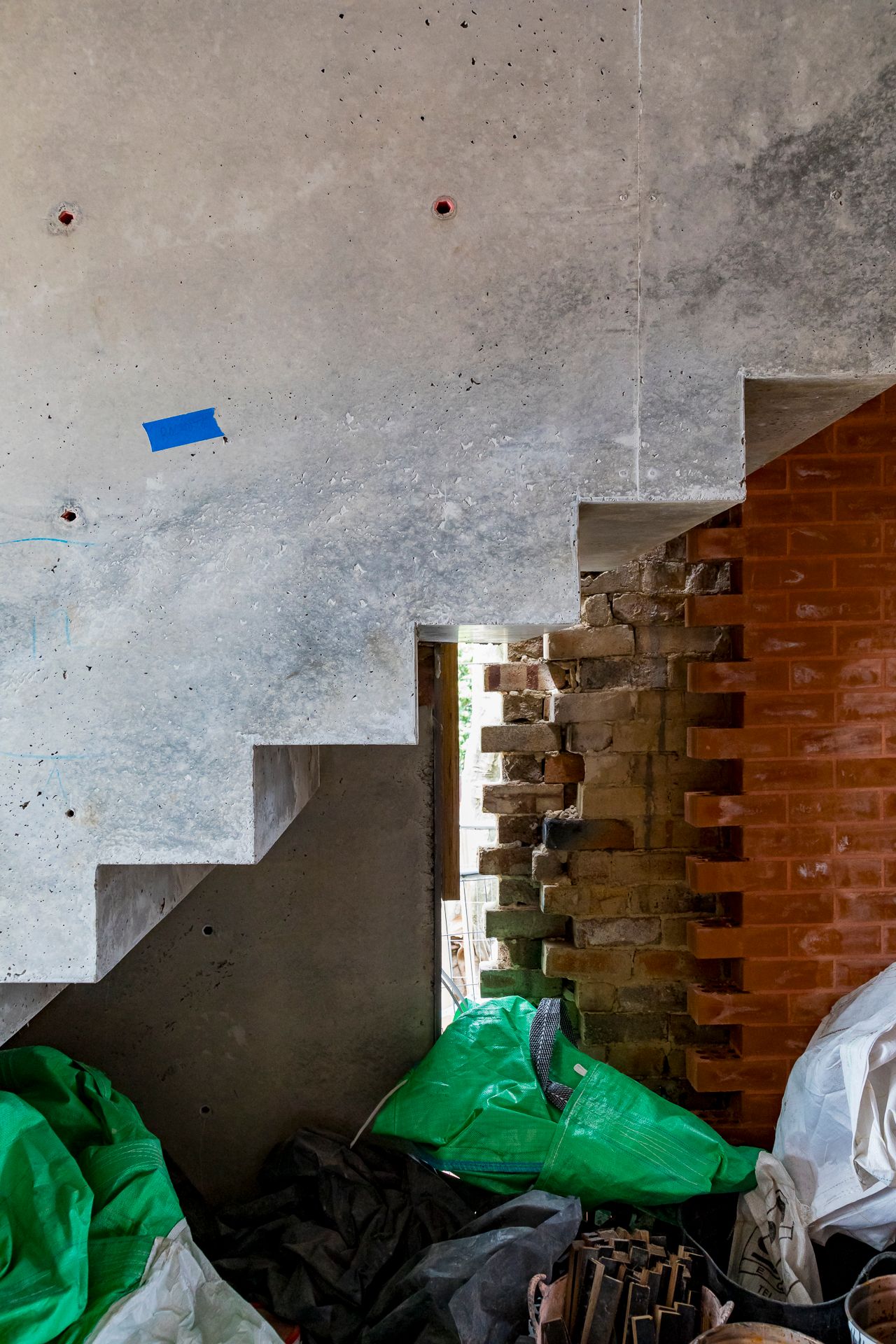 Raw concrete staircase underside with a blue marker tag beside exposed old brickwork, green tarps and site debris.