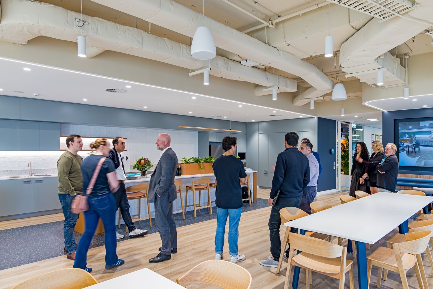 Team members gather in a bright office cafe area with white tables, wooden chairs and exposed ductwork overhead during a workplace tour.