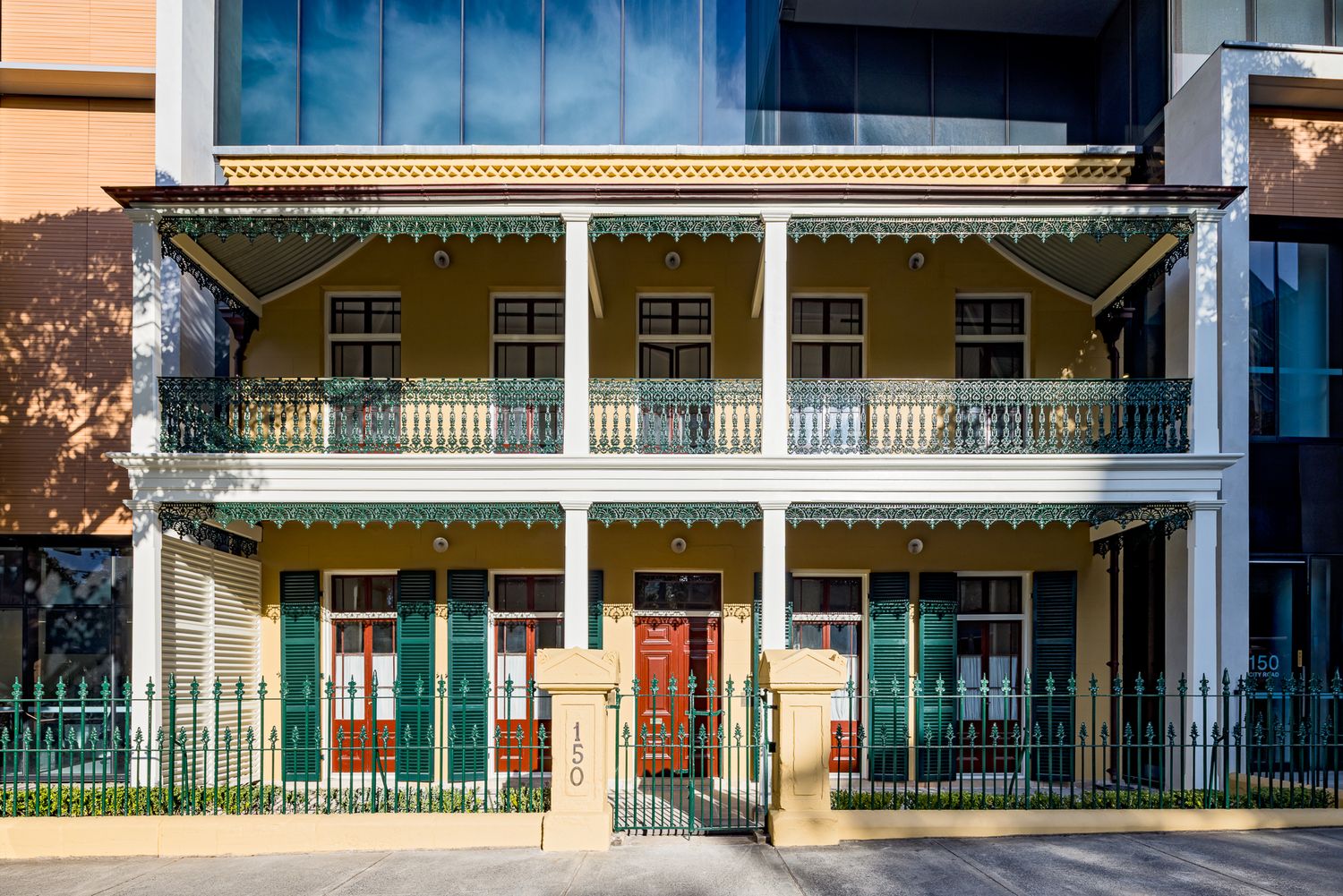 Front view of a heritage Victorian terrace with ornate green iron lacework balconies, red doors and a black iron fence.