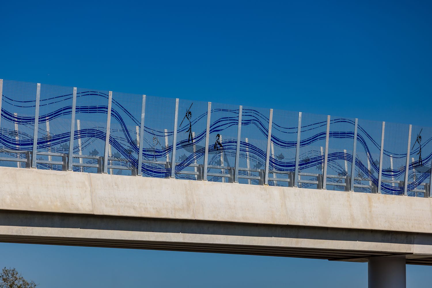 Close-up of a highway bridge barrier with artistic wave patterns and figure designs in blue glass panels against a blue sky.