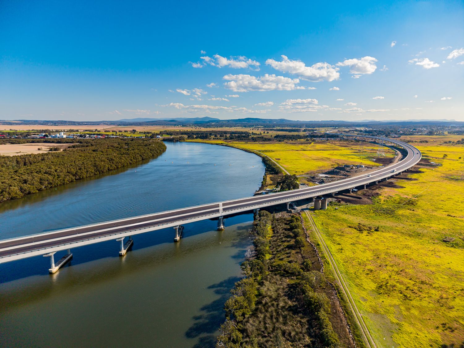 Drone view of a modern road bridge spanning a winding river beside yellow-green wetlands and distant hills under blue sky.