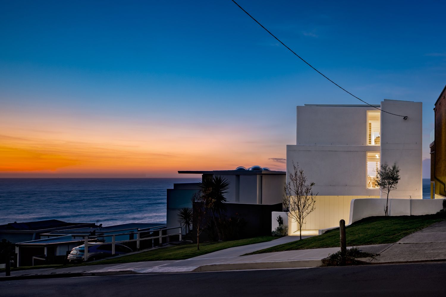 Modern white coastal house with lit windows overlooking the ocean at sunset beside a quiet seaside road.