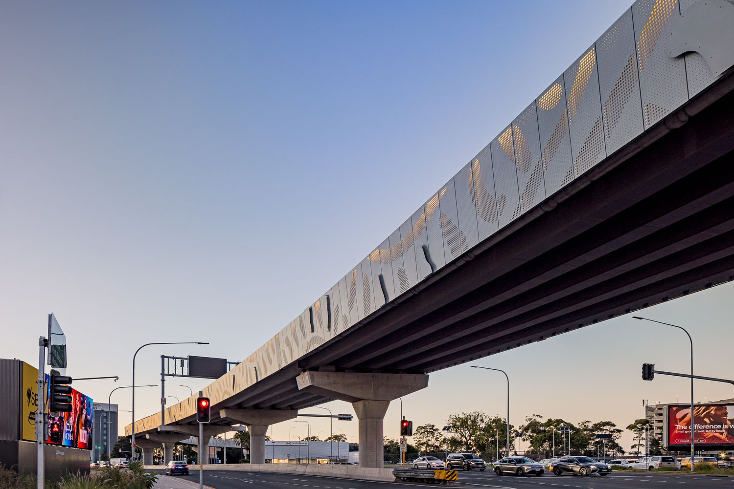 Low-angle view of a wide elevated transit bridge with wavy cladding and concrete supports above a busy intersection with traffic signals.