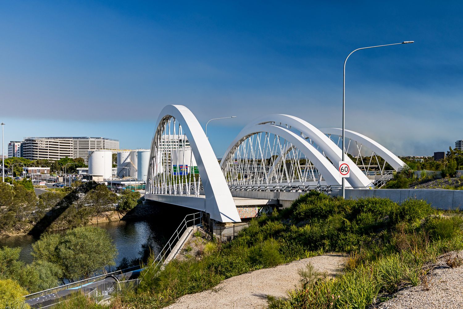 White steel arch bridge crossing a river with green embankments, industrial storage tanks and city buildings in the background.