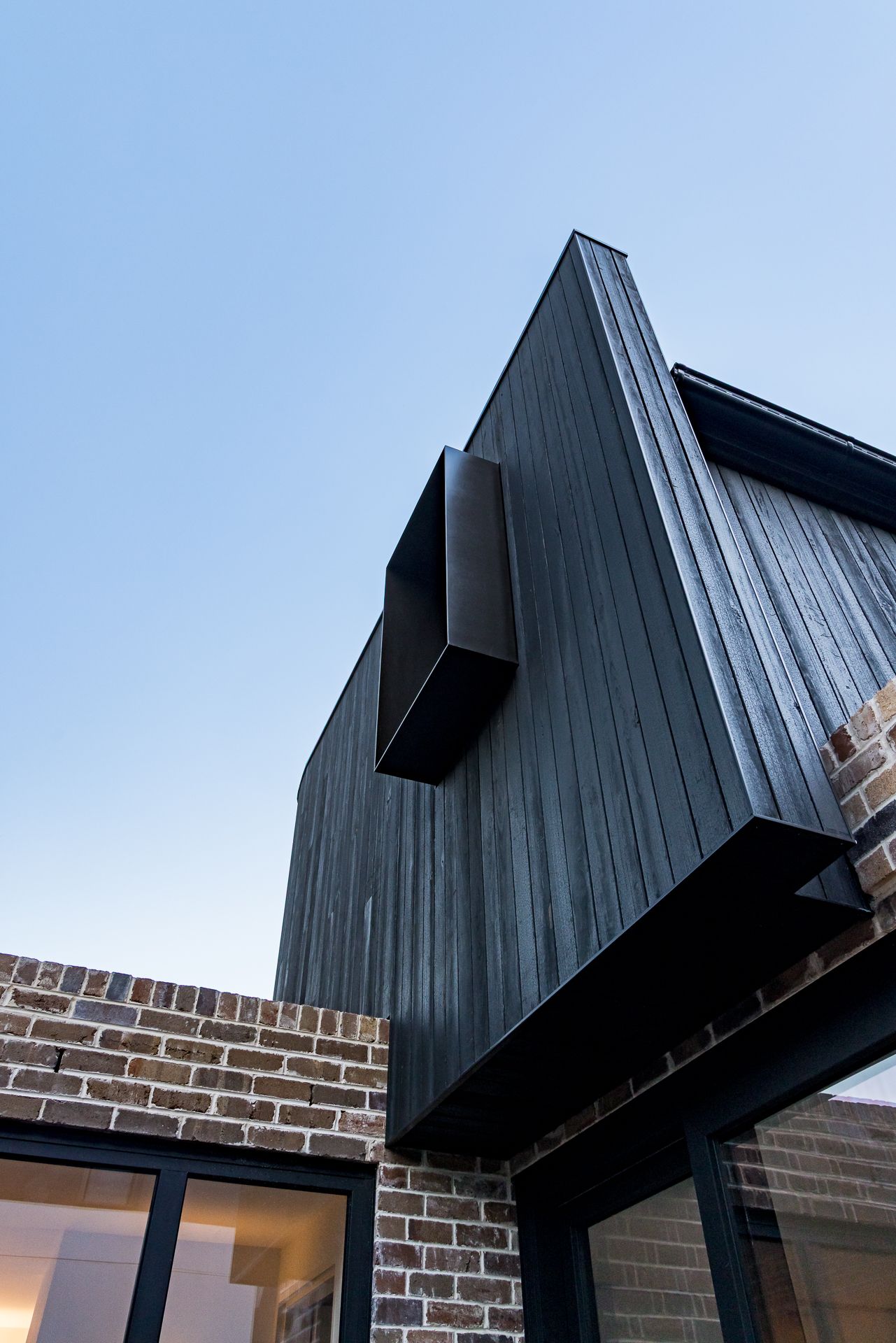 Dark charred timber cladding and a cantilevered window box rise above exposed brick walls against a clear dusk sky.
