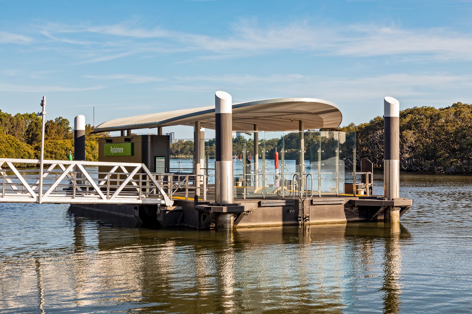 Small riverside ferry pontoon with a curved roof and white access ramp, set against bushland and a blue sky with light clouds.