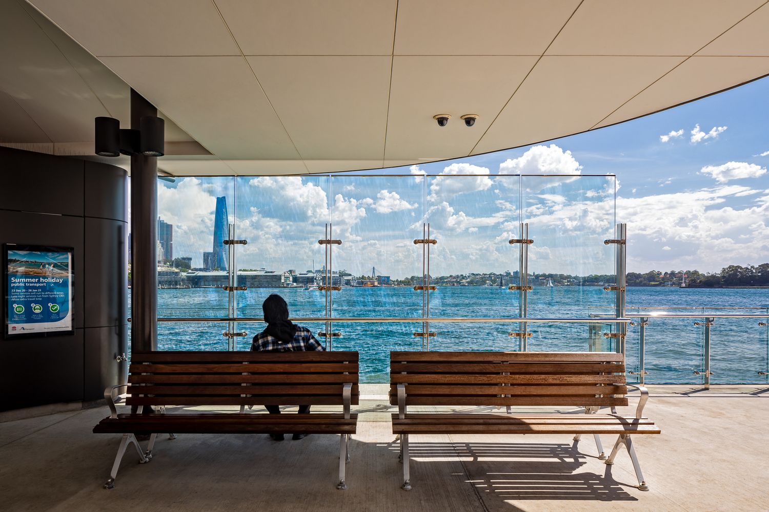 Person sits on a wooden bench in a covered ferry wharf, looking out over sparkling harbour water and a distant shoreline.