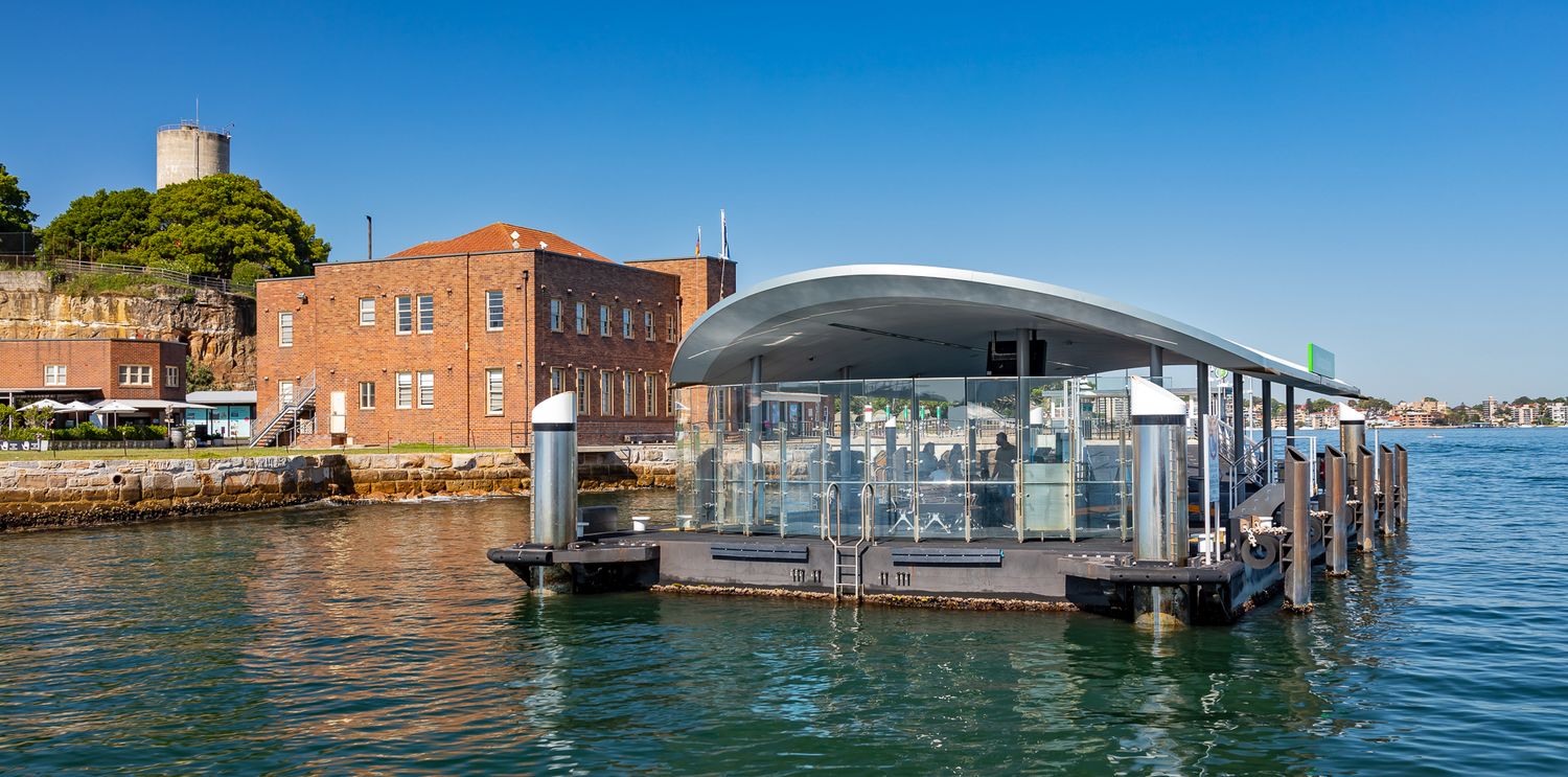 Curved-roof glass ferry terminal floating on harbour water, next to a historic red brick building and a stone water tower.