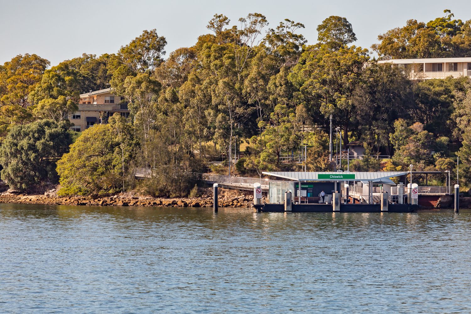 Small ferry wharf and pontoon on a calm harbour, backed by a rocky shoreline and dense bushland under a clear sky.