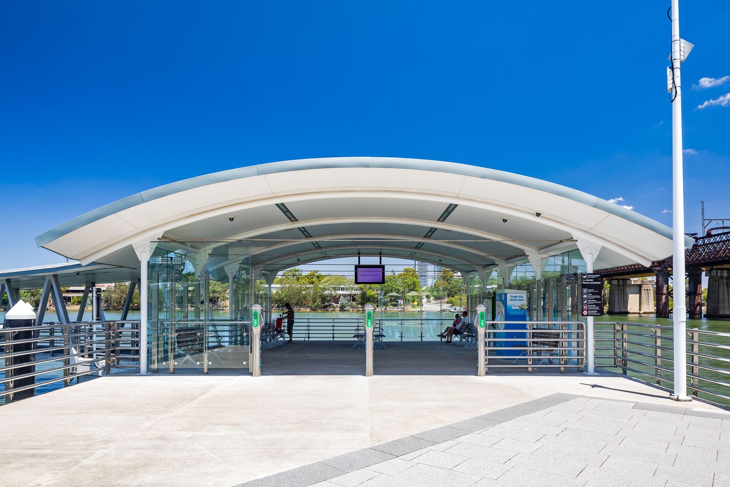 Modern curved-roof ferry terminal with glass walls and a wide paved walkway under a clear blue sky beside the river.