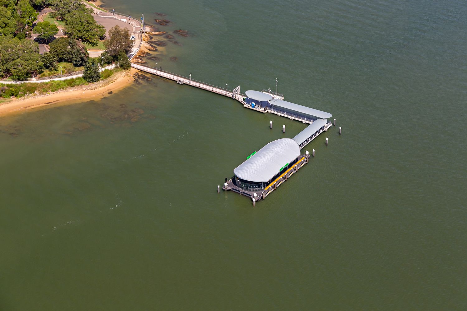 Aerial view of a long timber jetty leading to a curved-roof ferry wharf over calm green water, with a sandy shoreline and trees nearby.