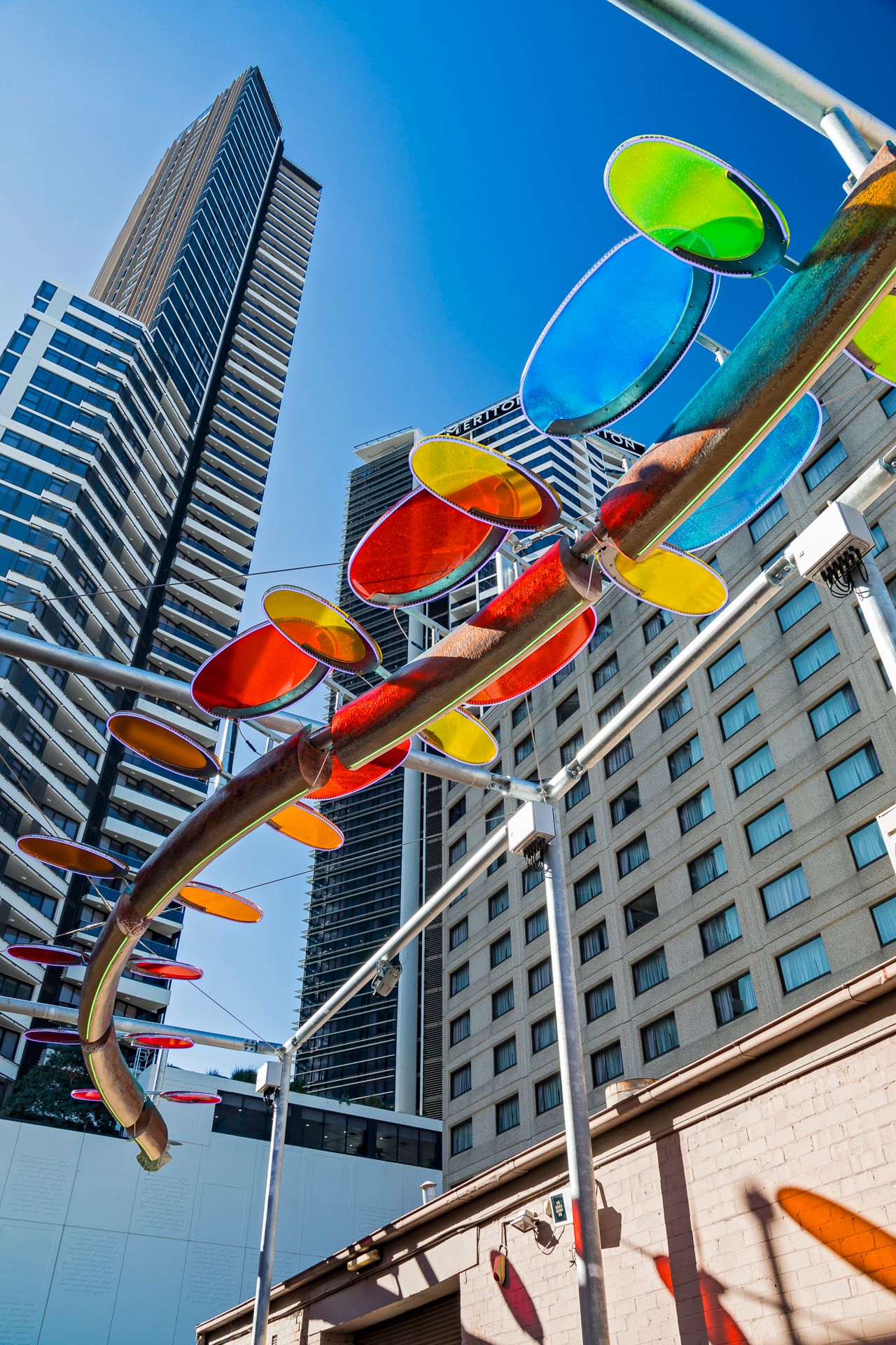 Colorful glass discs on a twisting copper sculpture overhead, with tall glass skyscrapers rising under a bright blue sky.