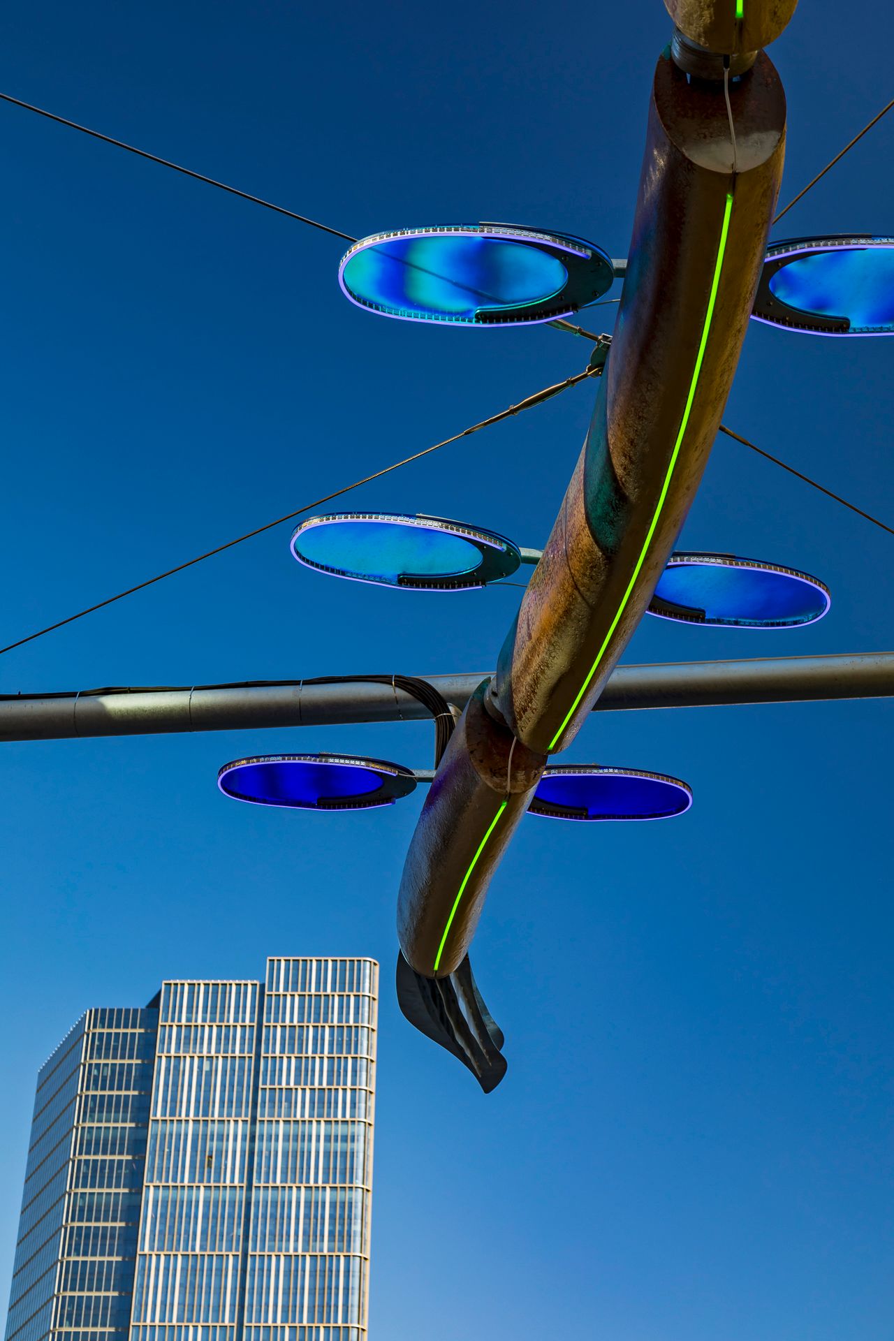 Low-angle view of a serpent sculpture with glowing blue oval discs and a green light line against a clear sky and glass tower.