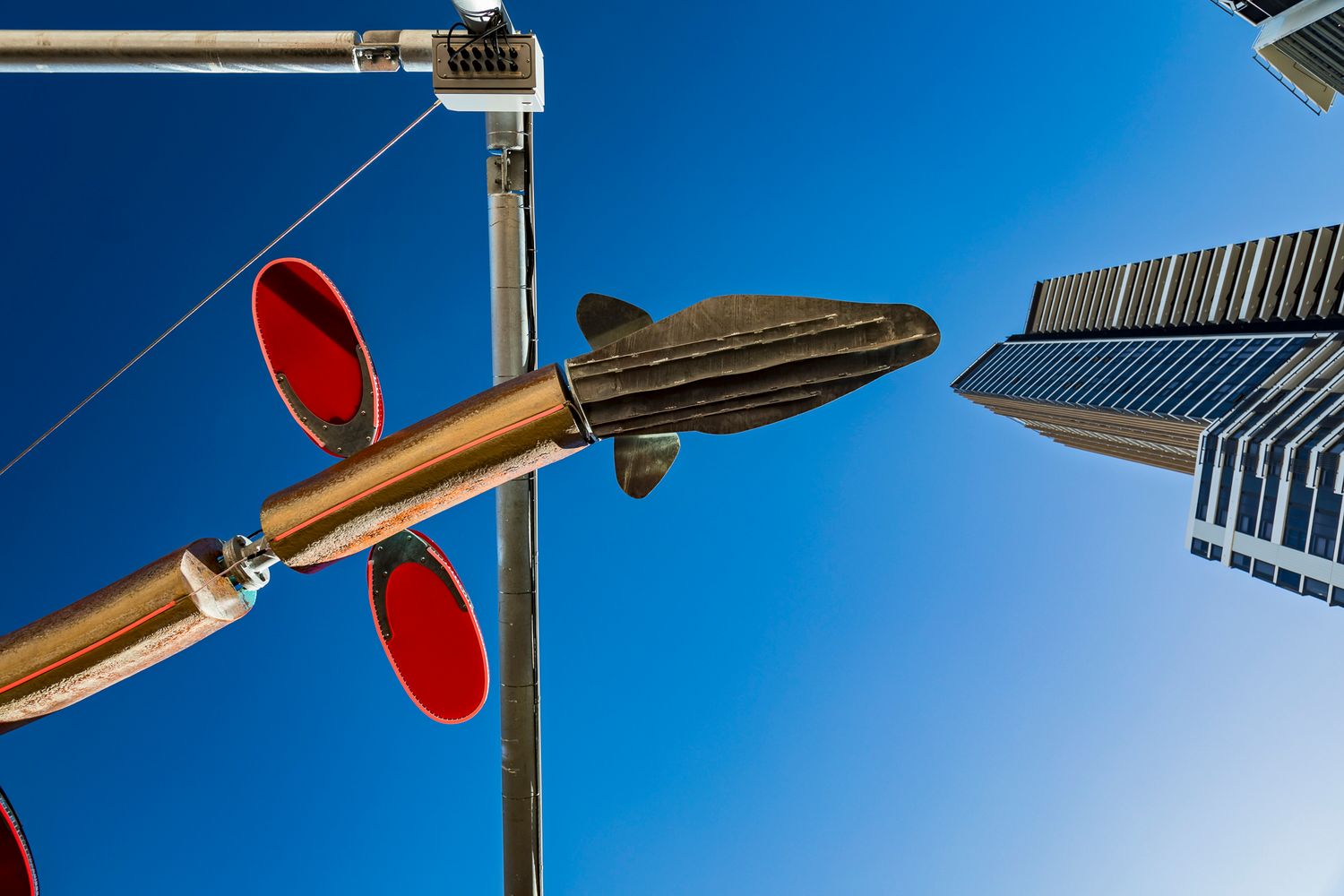 Upward view of a fish-like sculpture with red discs mounted on steel poles beside a modern high-rise tower.