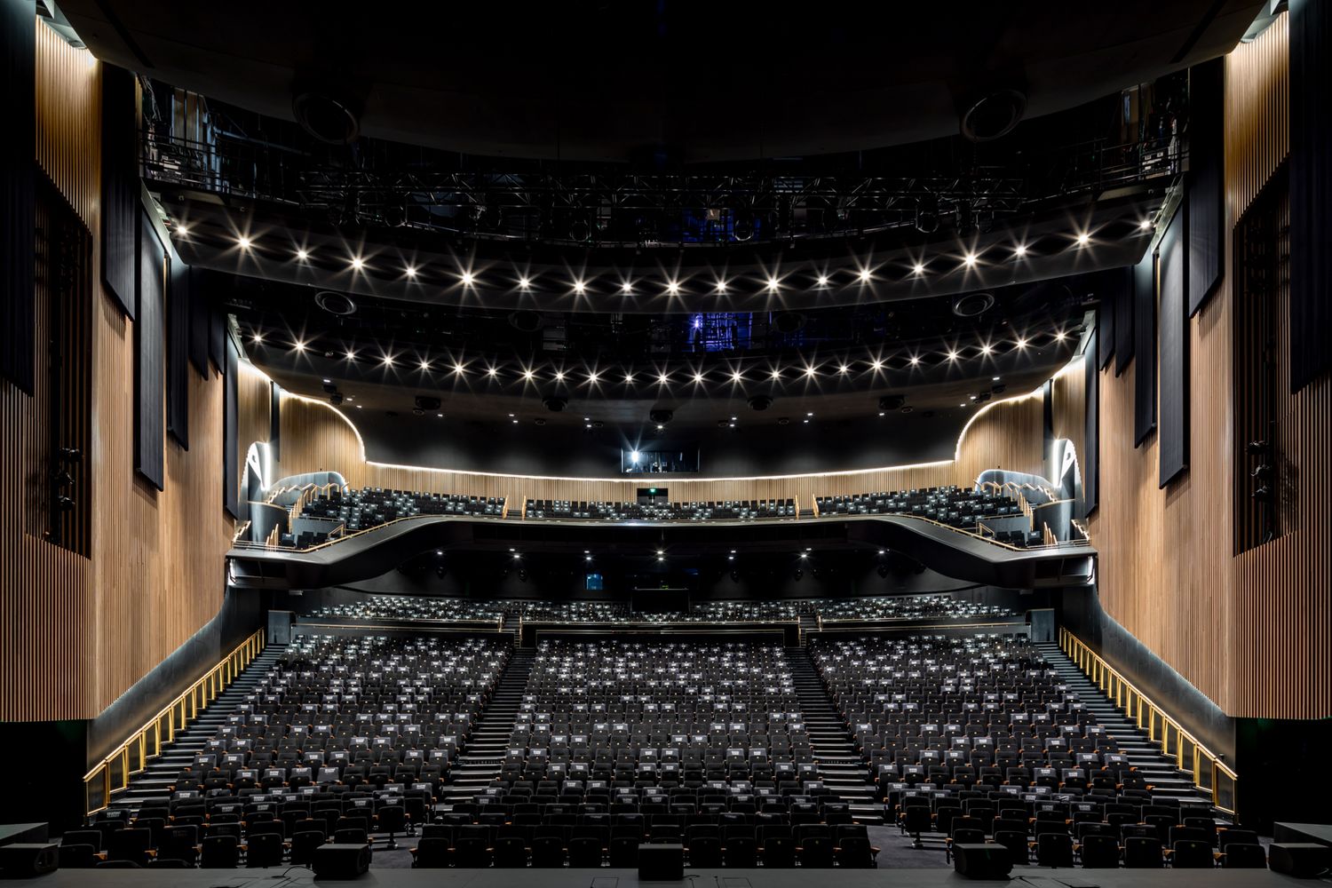 Spacious modern theatre auditorium with timber-clad walls, tiered seating and rows of ceiling lights above the stage.