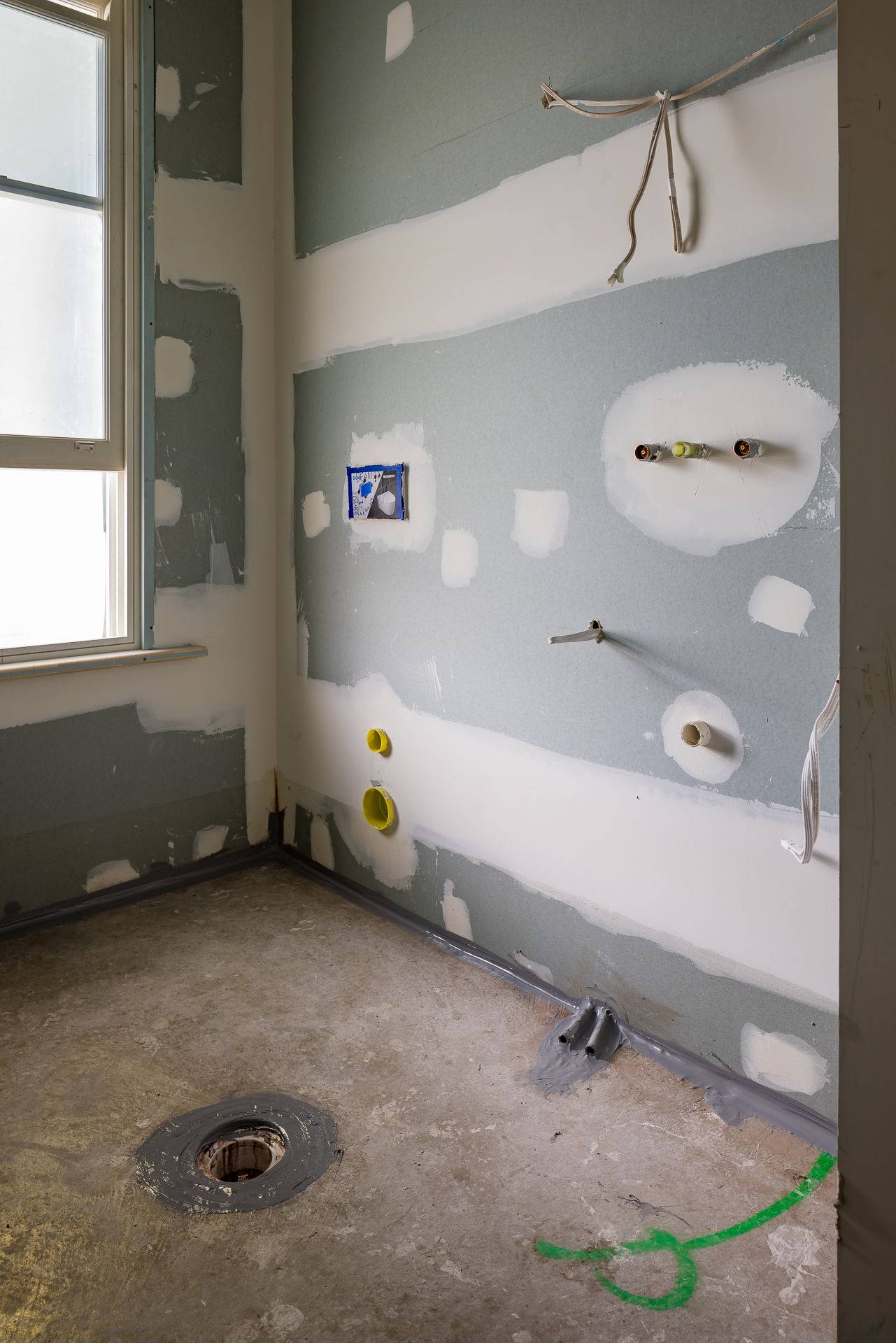 Bathroom under construction with exposed plumbing pipes, a floor drain and partially finished plasterboard walls beside a window.