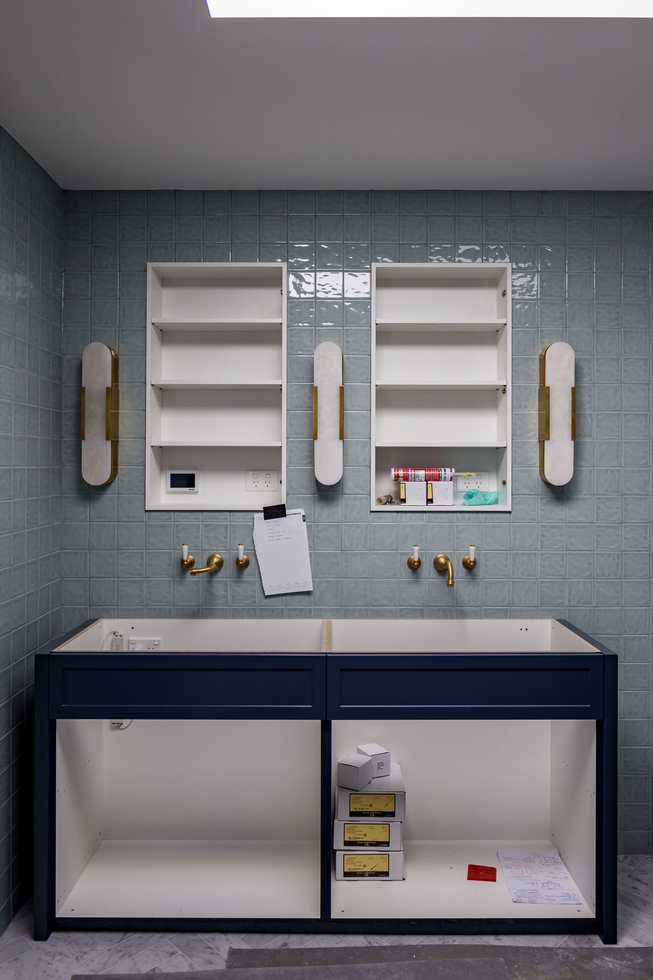 Bathroom under construction with sage green glazed tiles, brass wall taps, navy vanity cabinet, recessed shelves and wall sconces.