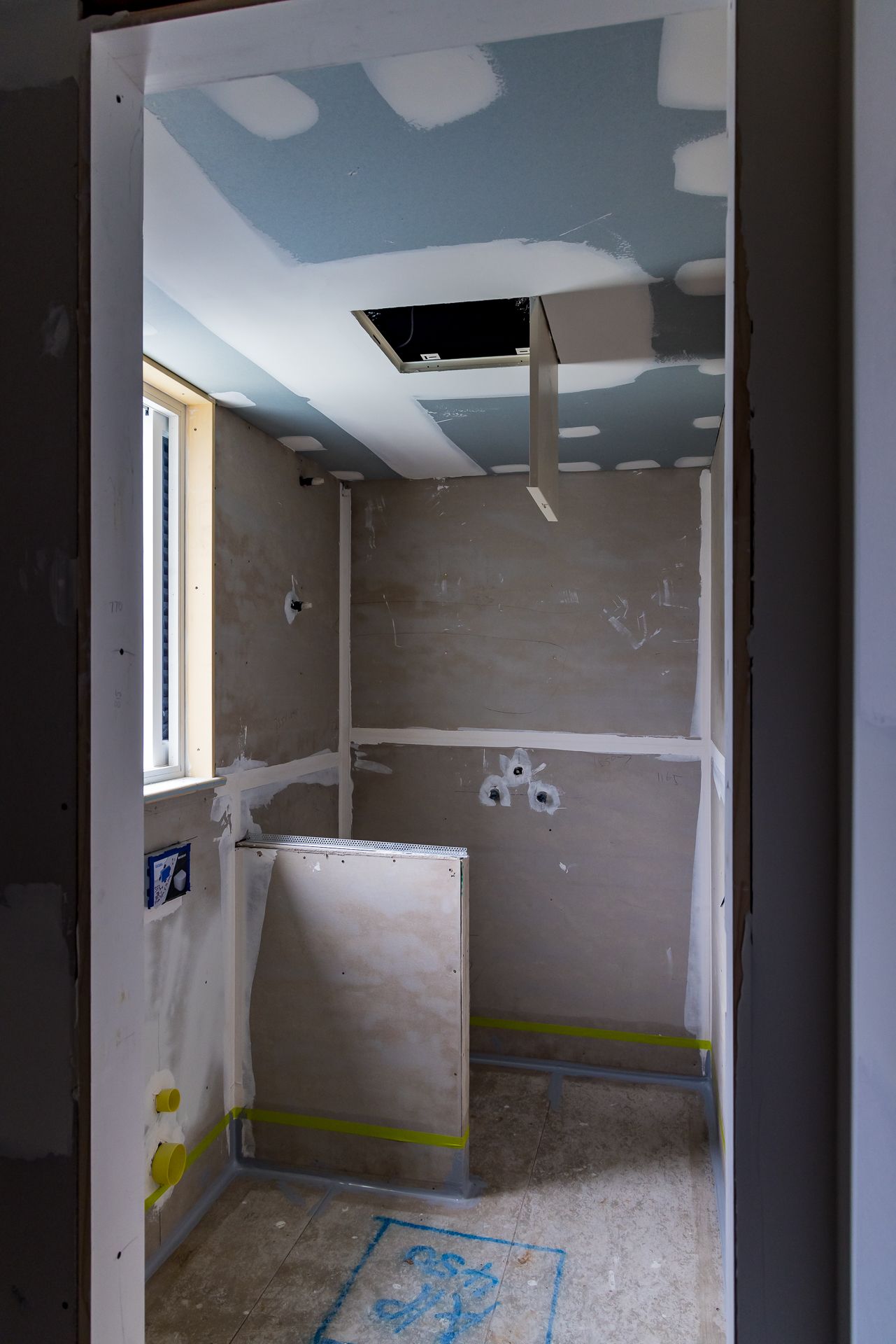 Unfinished bathroom under renovation with plasterboard walls, a blue-tinted ceiling, a small window and a ceiling access hatch.