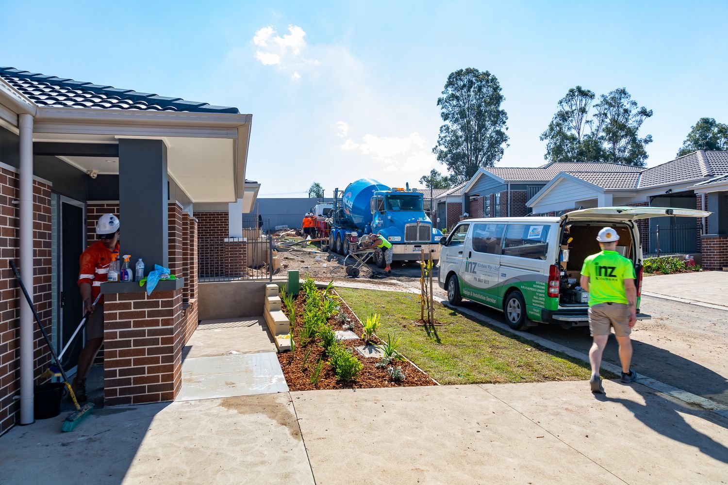 Construction workers in hi-vis gear and a white van at a new housing site with a blue excavator and landscaped front gardens.