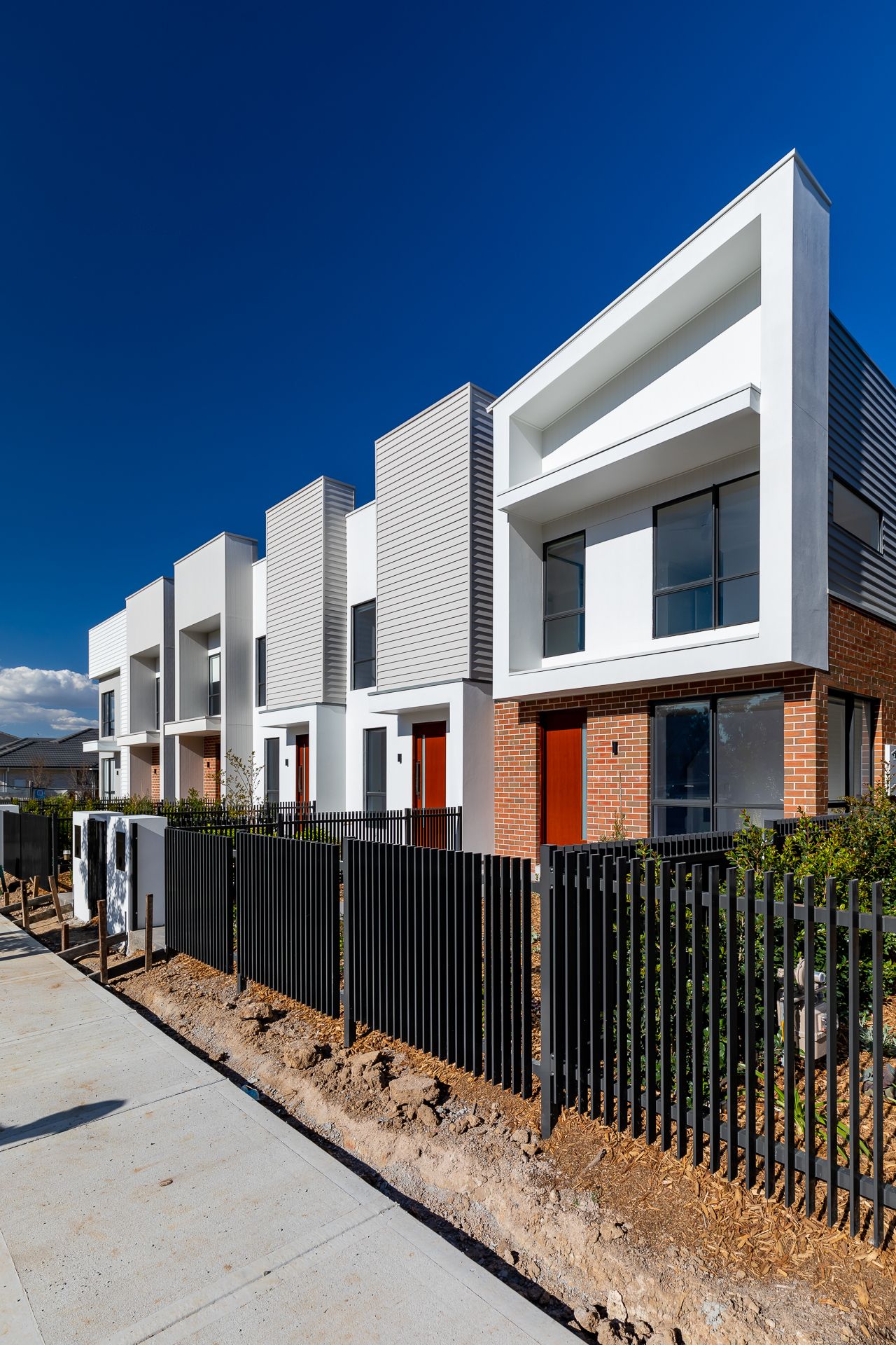 Row of modern white and grey townhouses with red front doors, brick accents and a black metal fence beside a footpath.