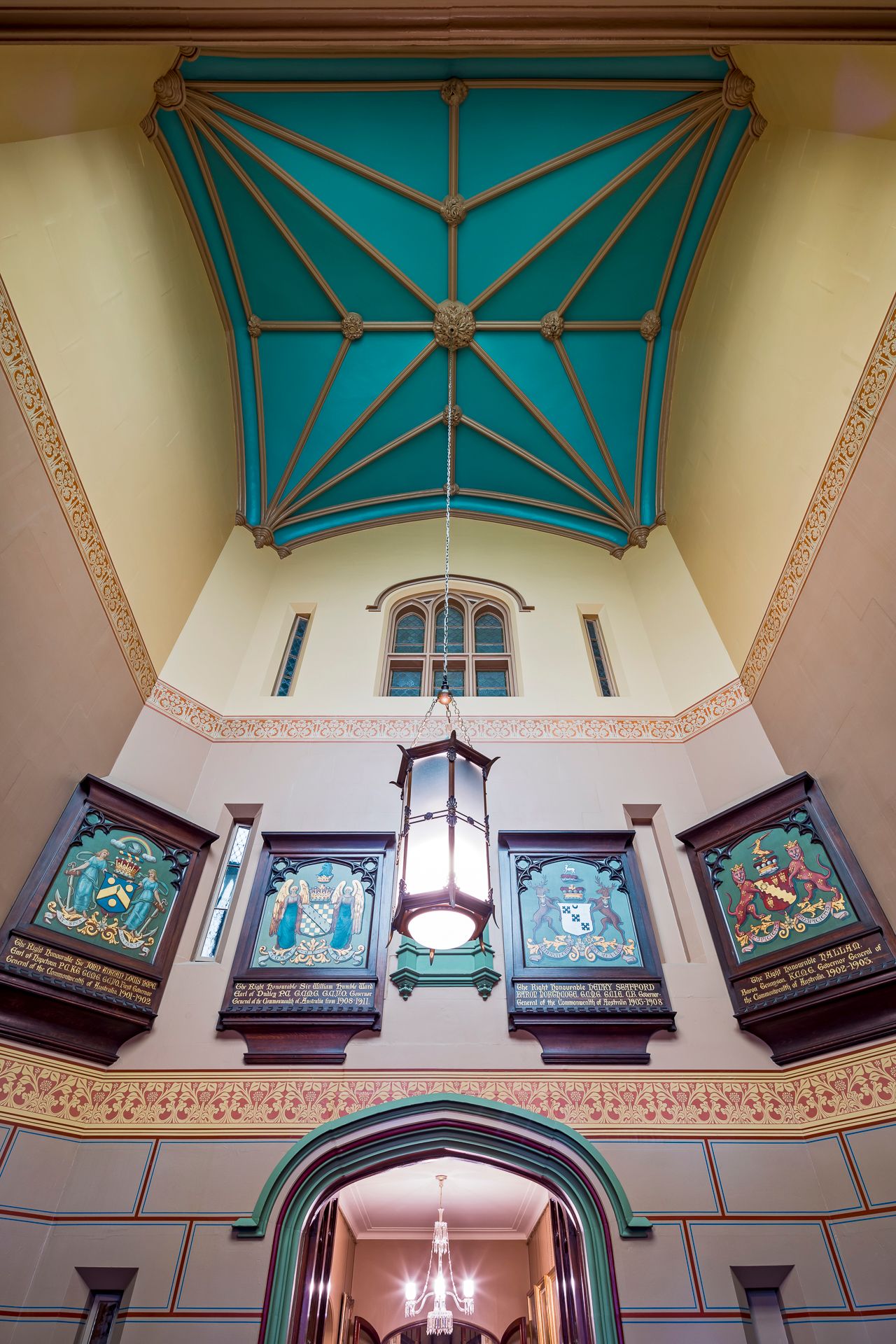 Heritage stairwell with a teal vaulted ceiling, hanging lantern, arched window and carved wooden coat of arms panels.