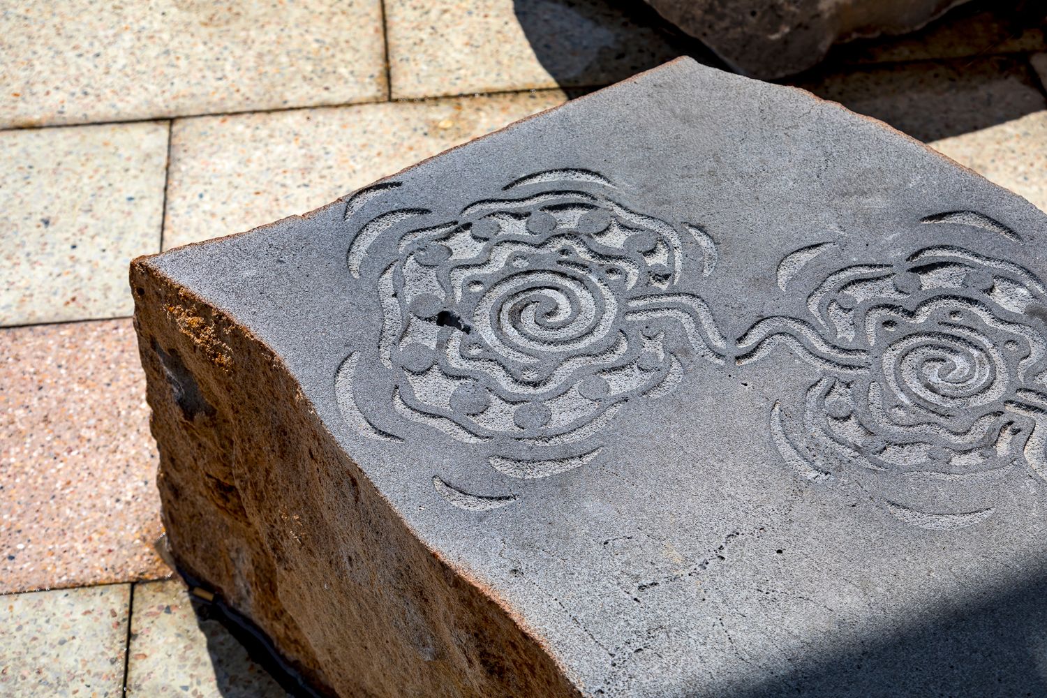 Close-up of a dark stone boulder with engraved spiral floral patterns resting on pale brick pavers.