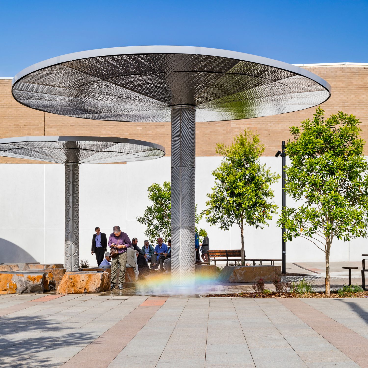 Circular metal shade canopies on slim columns tower over a paved plaza where people sit on rock seating beside young trees.