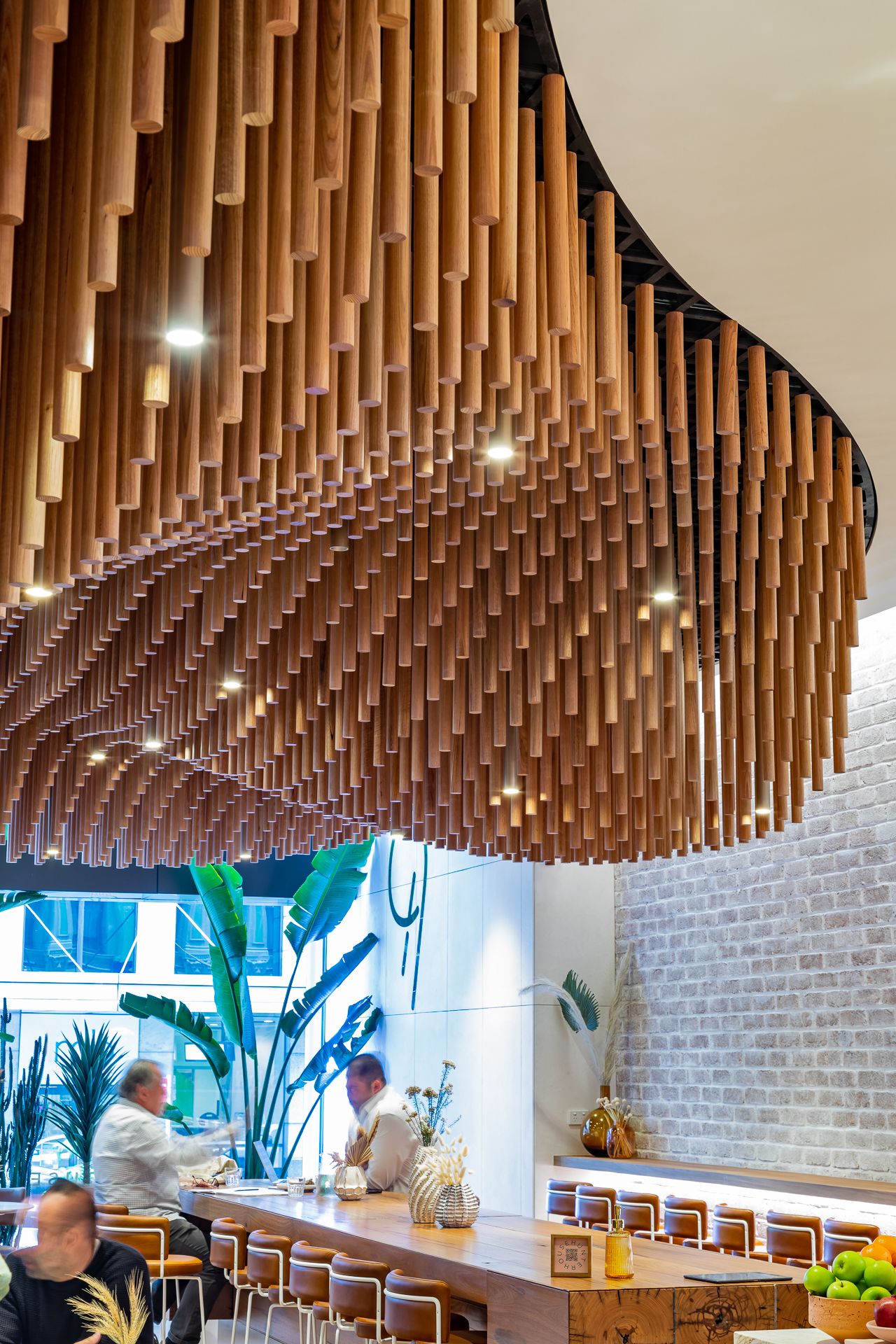 Cafe interior with a sculptural suspended ceiling of cascading timber tubes above a wooden counter, with patrons seated at the bar.