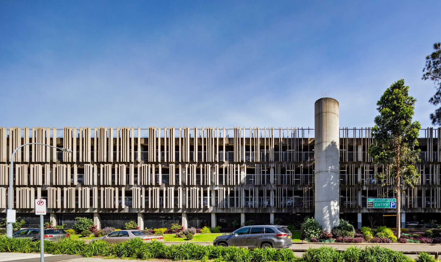 Modern multi-level car park with a tall concrete stair tower and a textured screen facade, framed by trees under a blue sky.