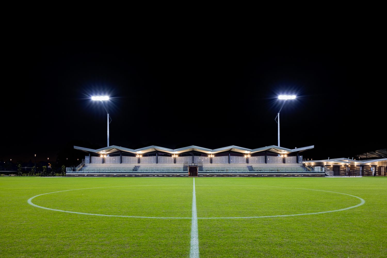 Floodlit football pitch at night with a covered grandstand, glowing stadium lights and a bright white center line on green turf.