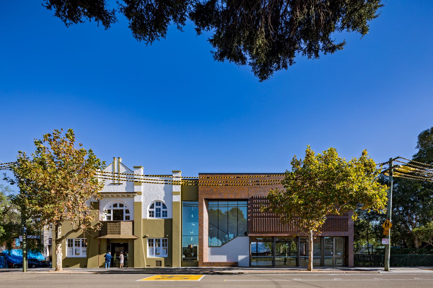 Heritage shopfront beside a modern brick and glass extension on a tree-lined street under a clear blue sky.