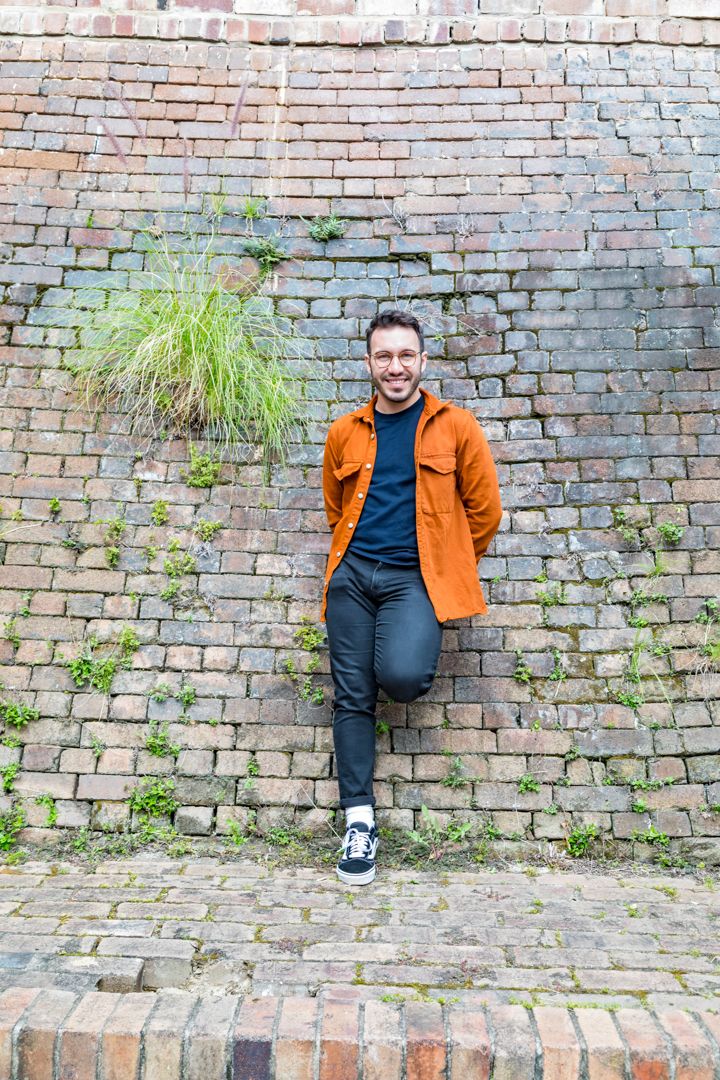 Smiling man in glasses, an orange jacket and black jeans leans against a weathered brick wall with grass growing from it.