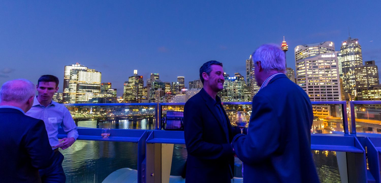 Two men in suits talk on a waterfront balcony at dusk, with glowing city skyline buildings and two other men standing nearby.