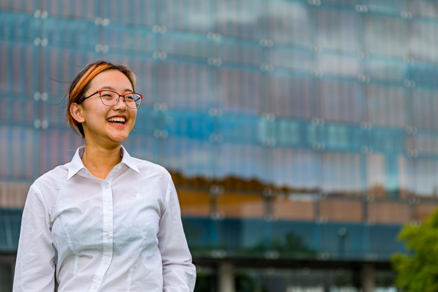 A smiling woman with glasses and a white collared shirt stands outdoors in front of a modern glass building.