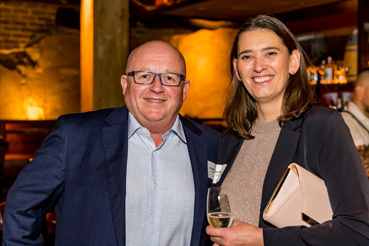 Smiling man in glasses and navy blazer stands beside a woman holding a glass of wine and a clutch at a warmly lit networking event.
