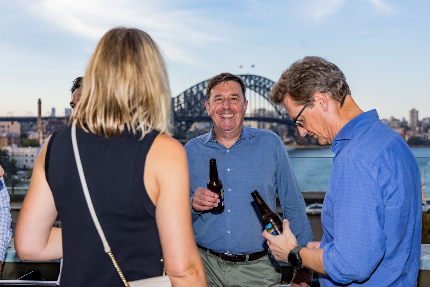 Smiling man in a blue shirt talks with two people holding beer bottles at a social event with Sydney Harbour Bridge in the background.