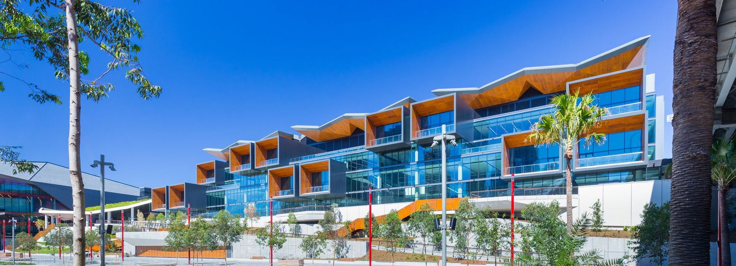 Modern multi-storey building with glass balconies and an orange angular roofline set among trees under a clear blue sky.