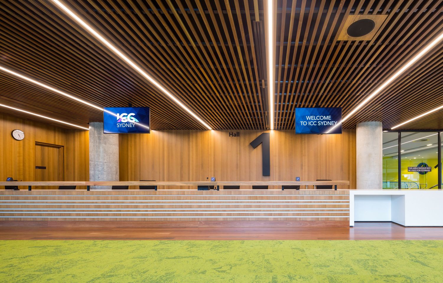 ICC Sydney welcome area with timber slat ceiling and wall panels, digital screens, reception counter and bright green carpet.