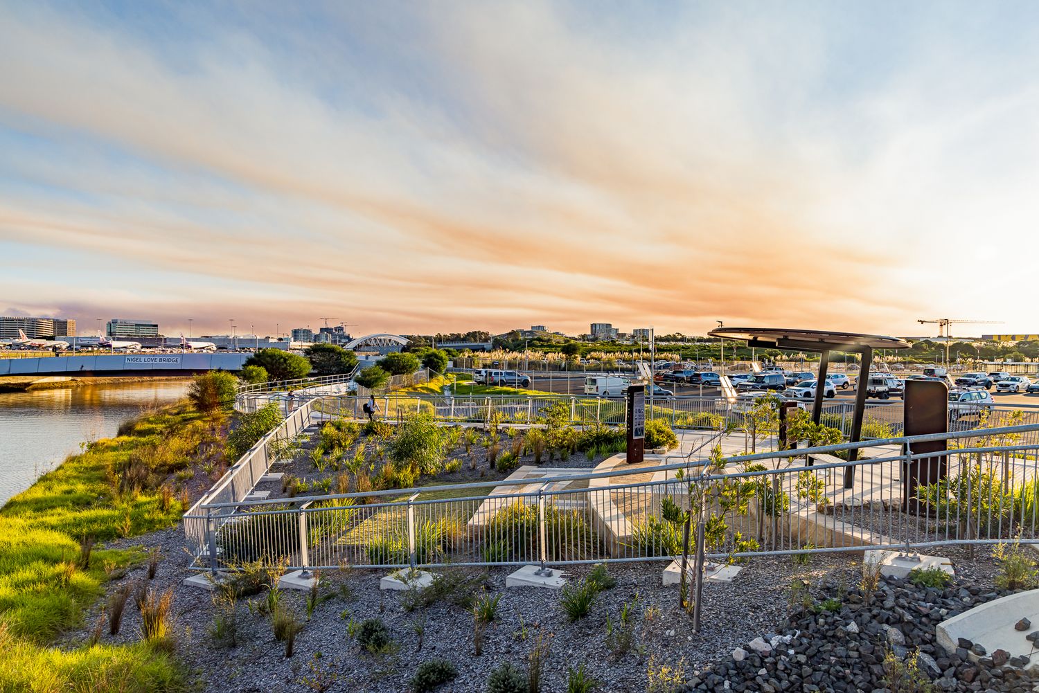 Sunset sky over a waterfront park with native plantings, winding paved paths, metal railings and distant city buildings.
