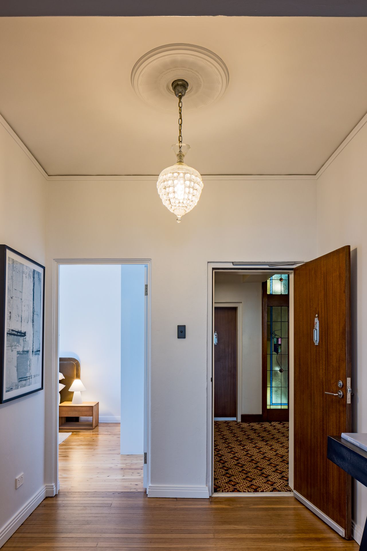 Bright apartment hallway with crystal pendant light, ceiling rose, timber floors and open doors to a bedroom and entry.