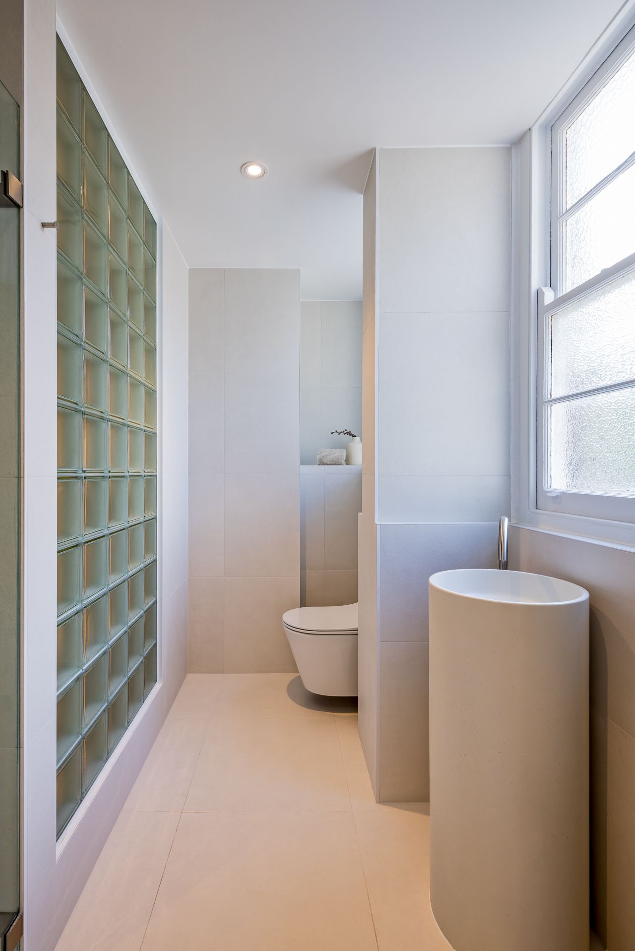 Minimalist bathroom with cream tiles, glass block wall, wall-mounted toilet and a freestanding round pedestal basin by a window.